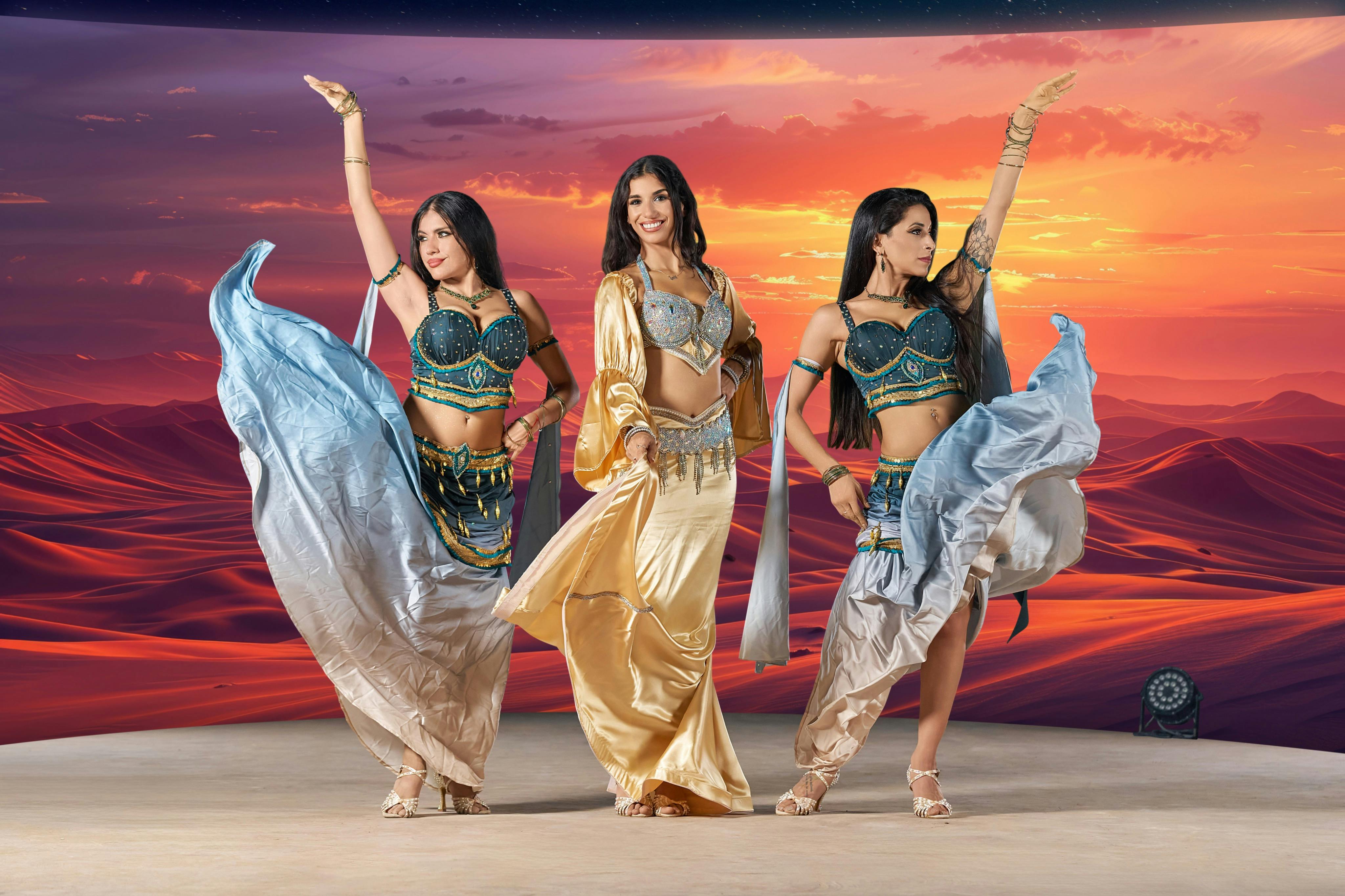 Three women in belly dance costumes pose against a vibrant sunset backdrop, with flowing skirts and raised arms.
