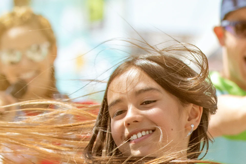 A girl dances joyfully with her hair whipping around, while four adults behind her sing enthusiastically in an outdoor setting with palm trees.