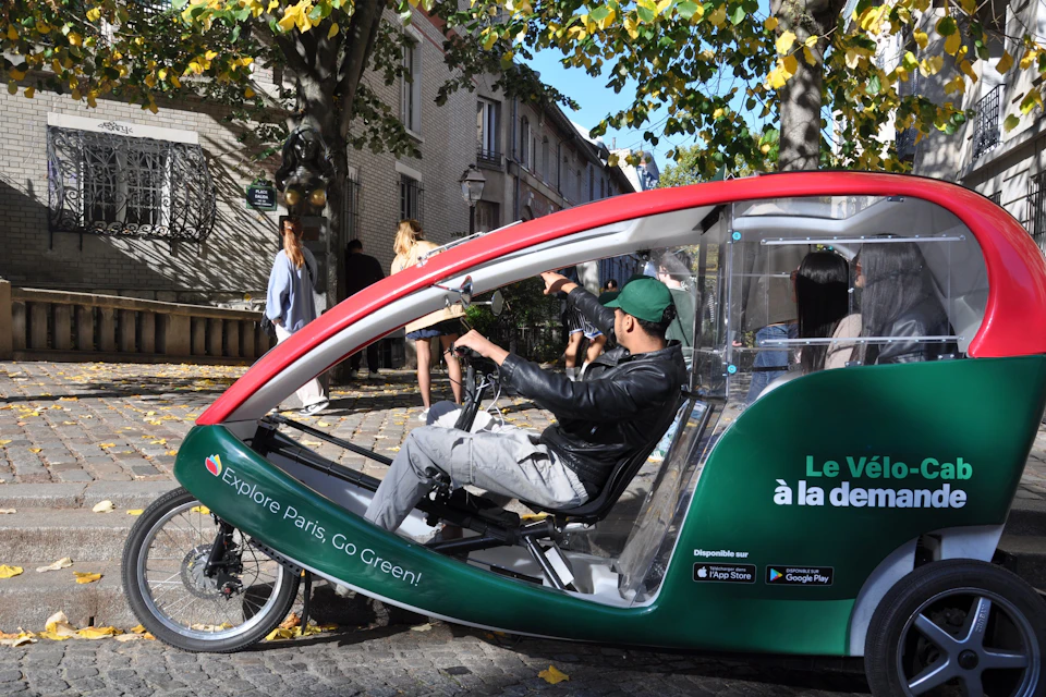 Person riding a green and red pedicab with "Explore Paris. Go Green!" written on the side, on a tree-lined street.