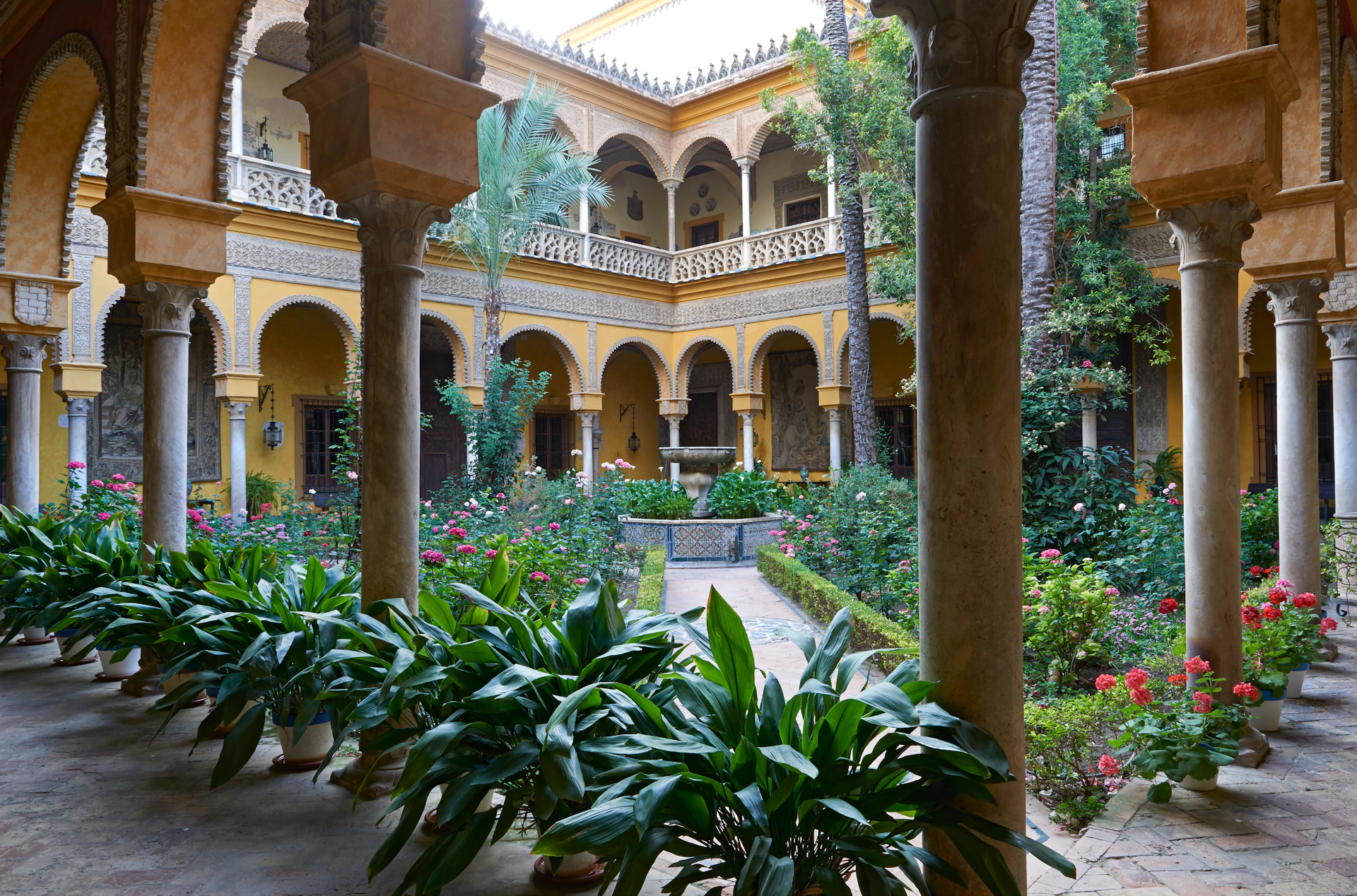 A courtyard with lush greenery, potted plants, arched yellow buildings, and a central fountain surrounded by flowers.