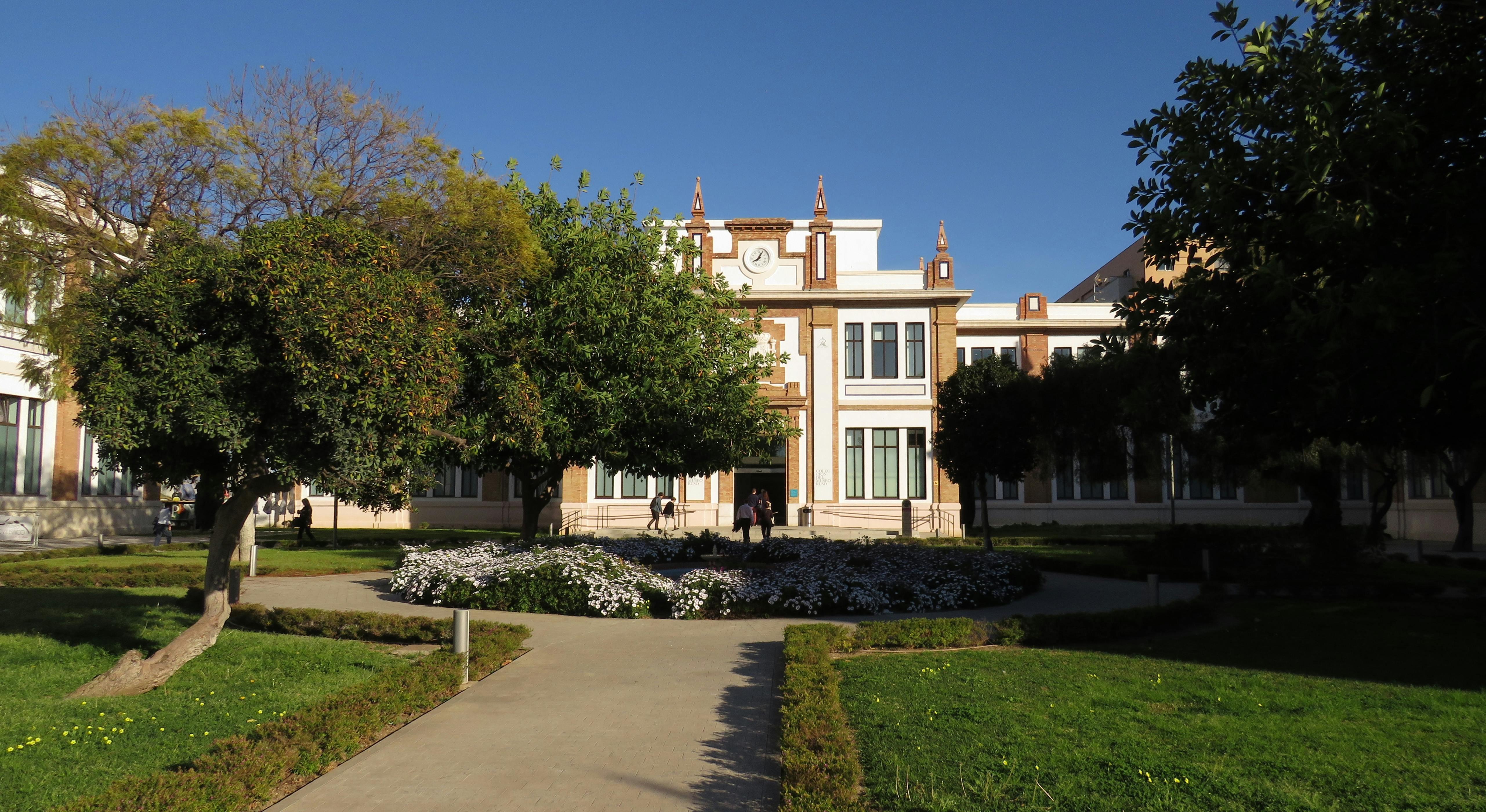 A building with a clock tower, surrounded by trees, flowers, and walking paths, under a clear blue sky. People near the entrance.