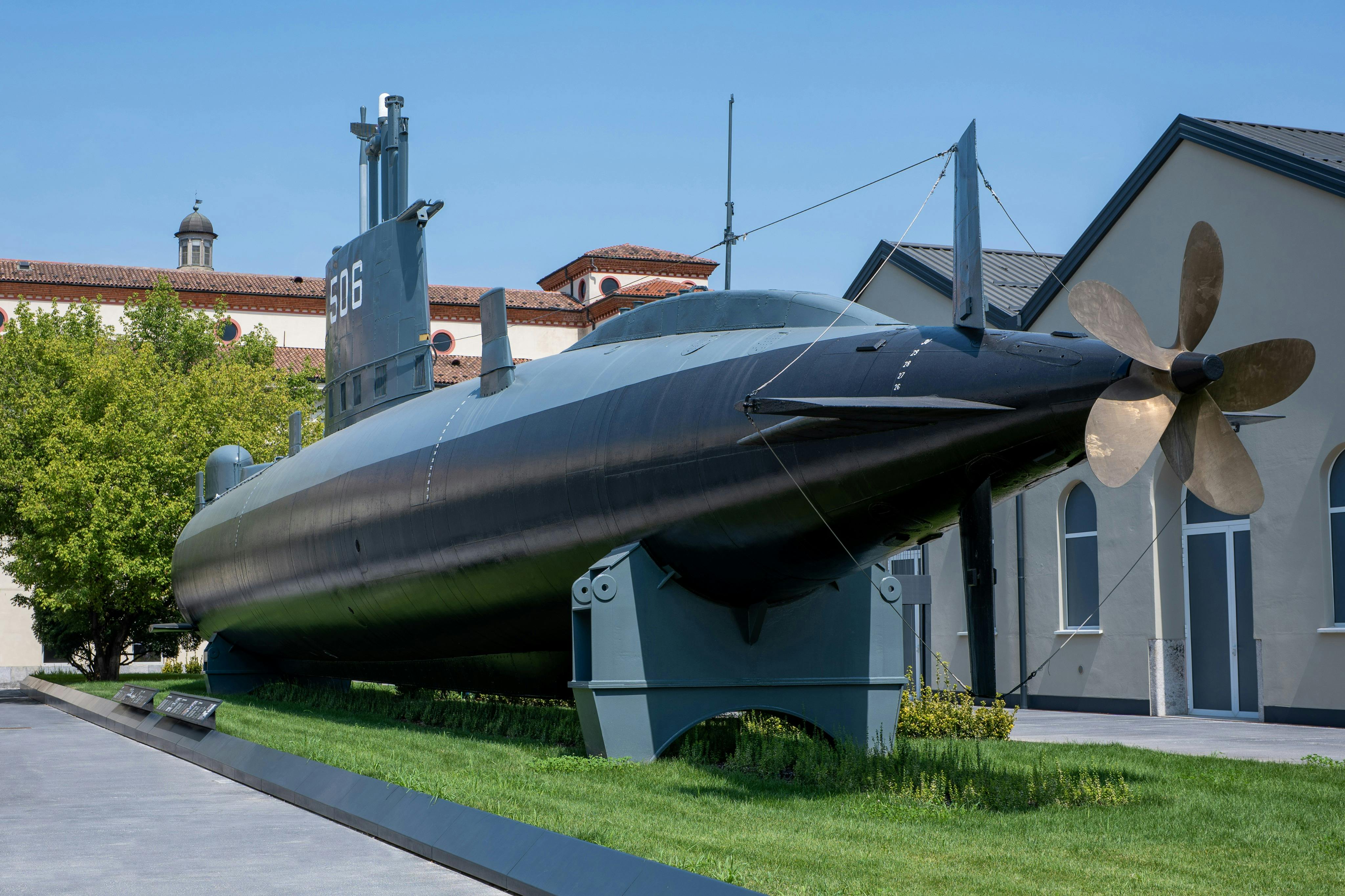 Un sottomarino nero esposto su una piattaforma all’aperto vicino ad alcuni edifici, sotto un cielo limpido e azzurro.