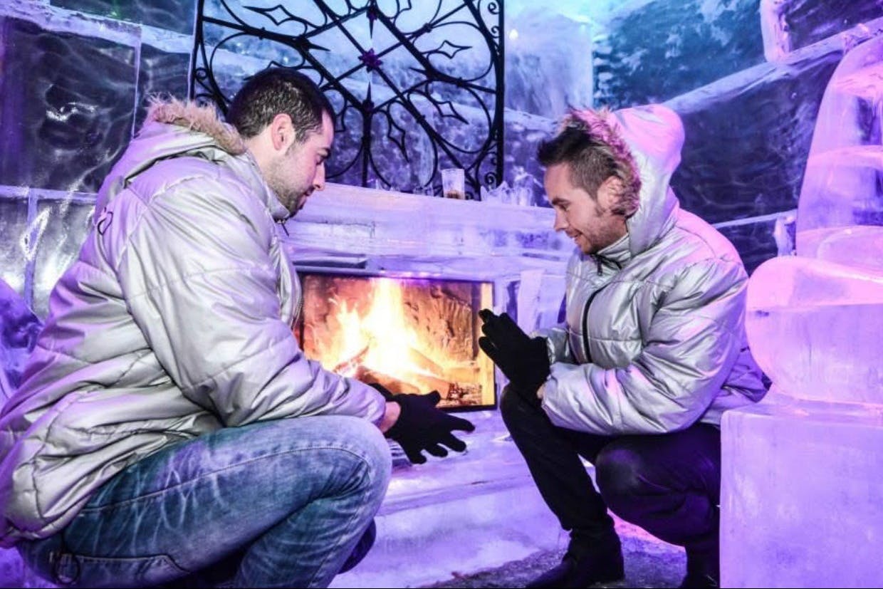 Deux personnes vêtues de vestes à capuche se réchauffent les mains près d'une cheminée dans un environnement glacial, avec un éclairage bleu et un décor de glace.