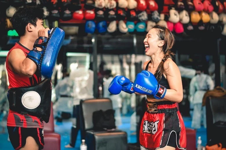 A woman wearing boxing gloves and red shorts smiles and laughs in a gym with boxing equipment and people in the background.