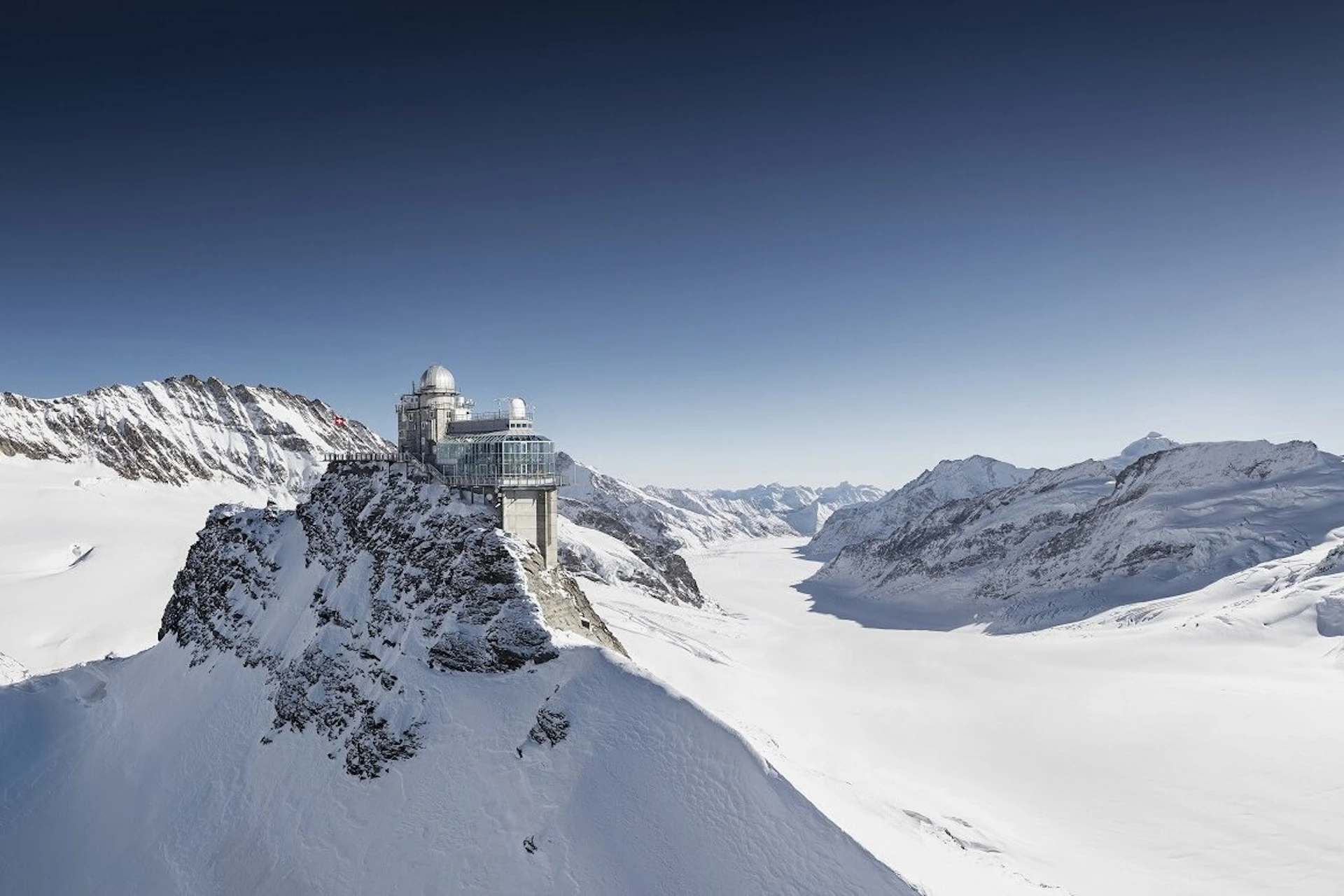Observatory and building complex atop a snowy mountain peak, surrounded by a vast alpine landscape under a clear blue sky.