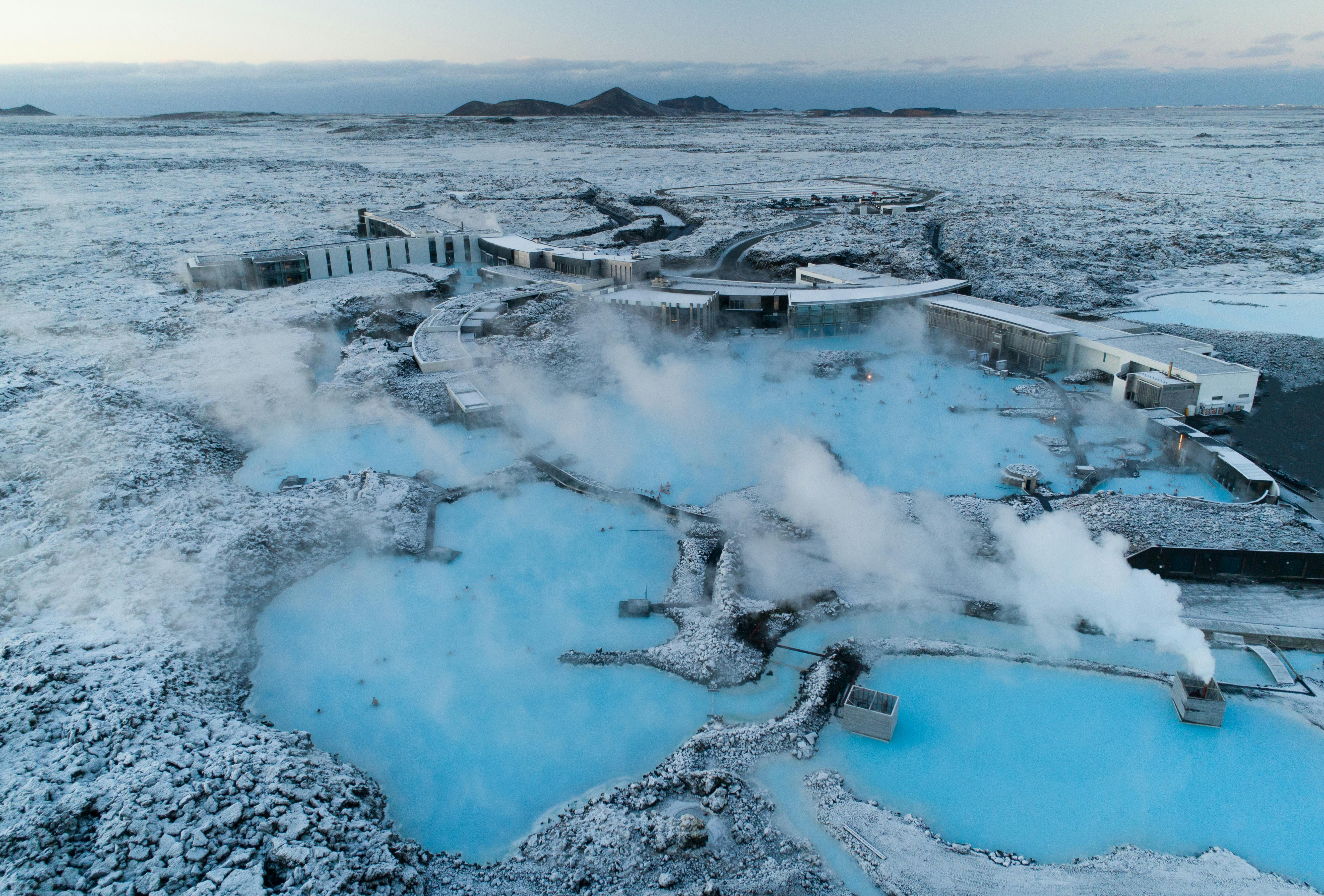 Veduta aerea di un centro termale geotermico con piscine di acqua blu fumante circondate da un terreno roccioso innevato e da edifici moderni.