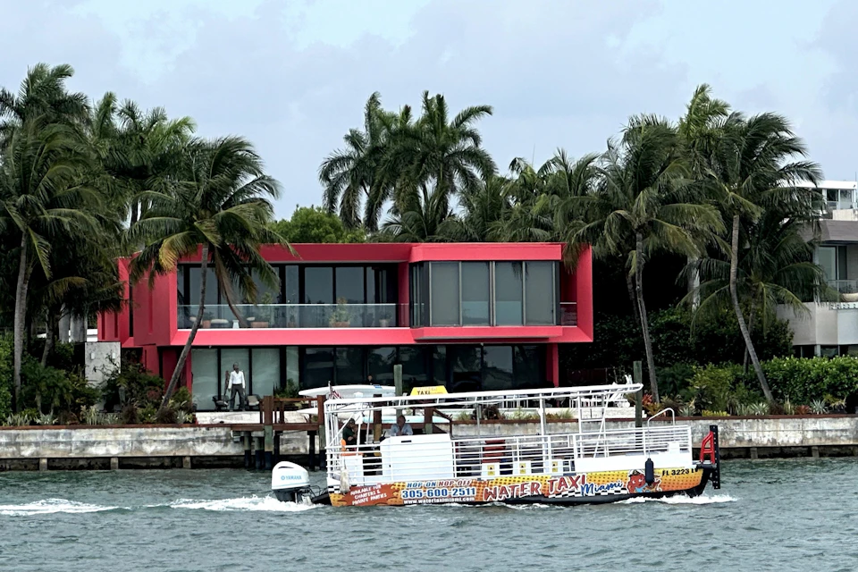 A water taxi passes in front of a modern red house with large windows and palm trees by the waterfront.