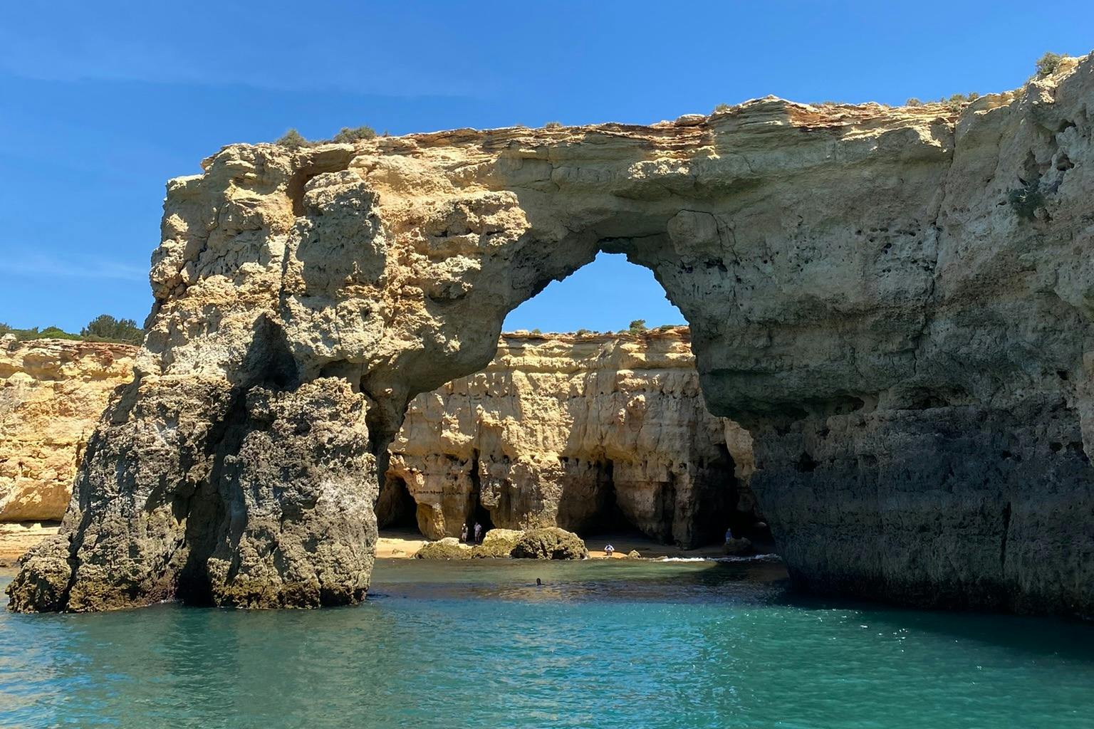 Une formation rocheuse en forme d'arche avec une petite plage et de l'eau bleue claire en dessous, sur fond de ciel bleu vif.