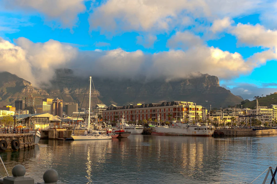 A harbor with boats docked in calm water, a red brick building, and a mountain partially covered by clouds in the background.