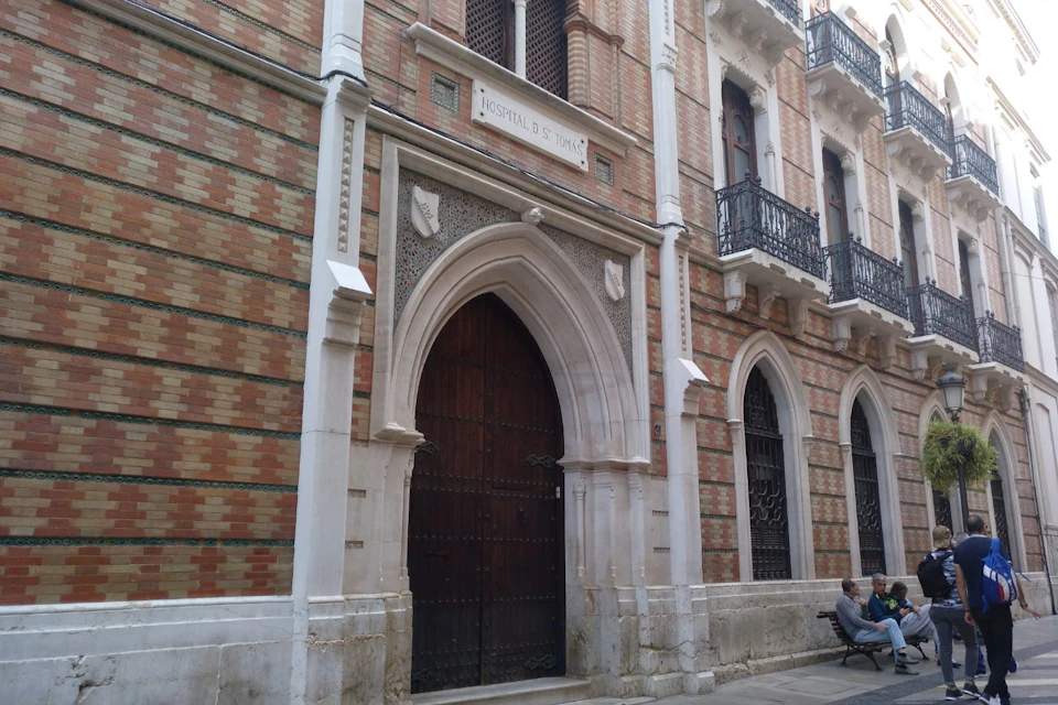 A brick building with an arched wooden door labeled "Hospital de S. Tomas," flanked by two windows and a person sitting nearby.