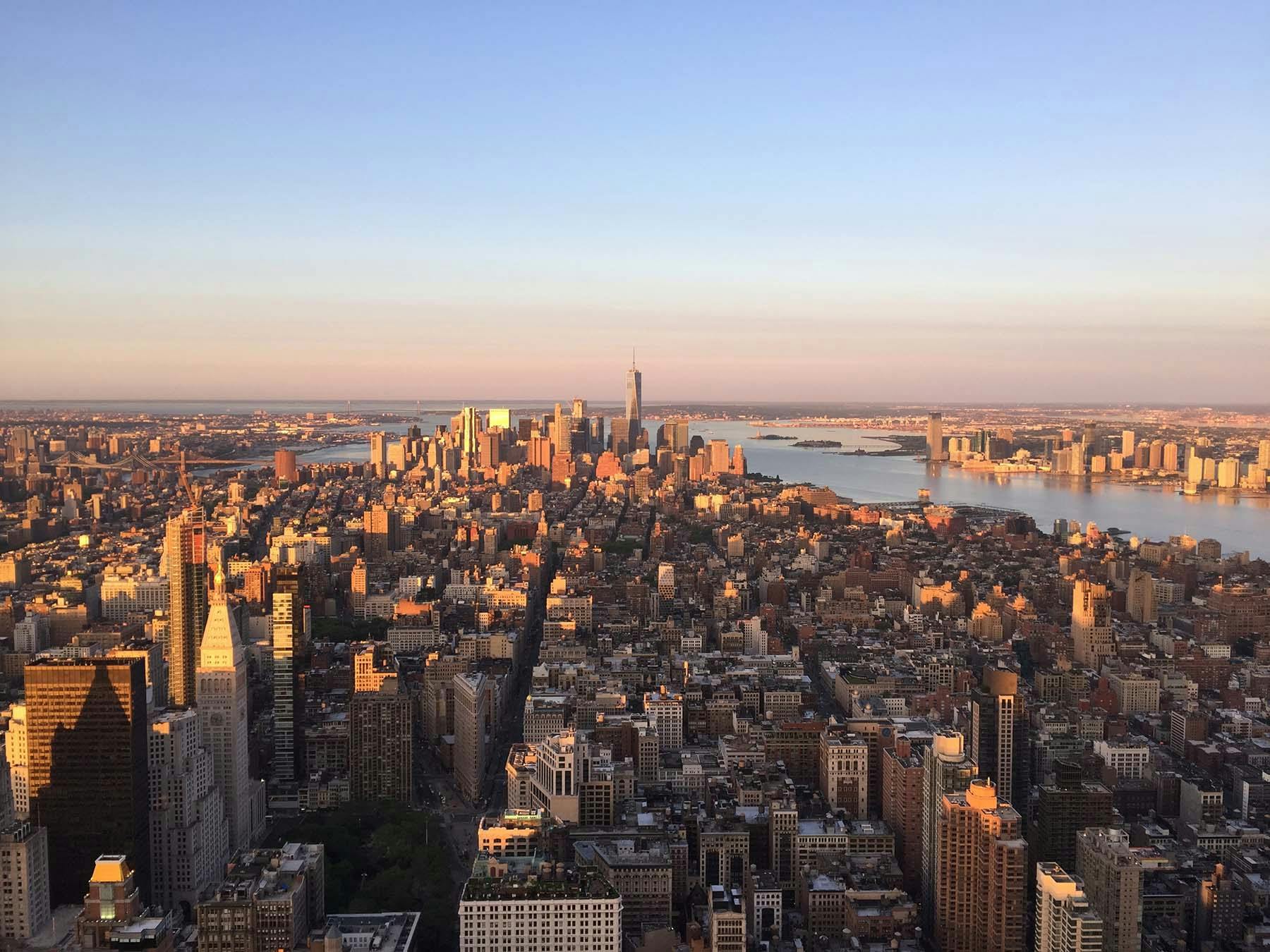 Aerial view of a densely packed cityscape at sunset with tall buildings, a river, and a clear sky.