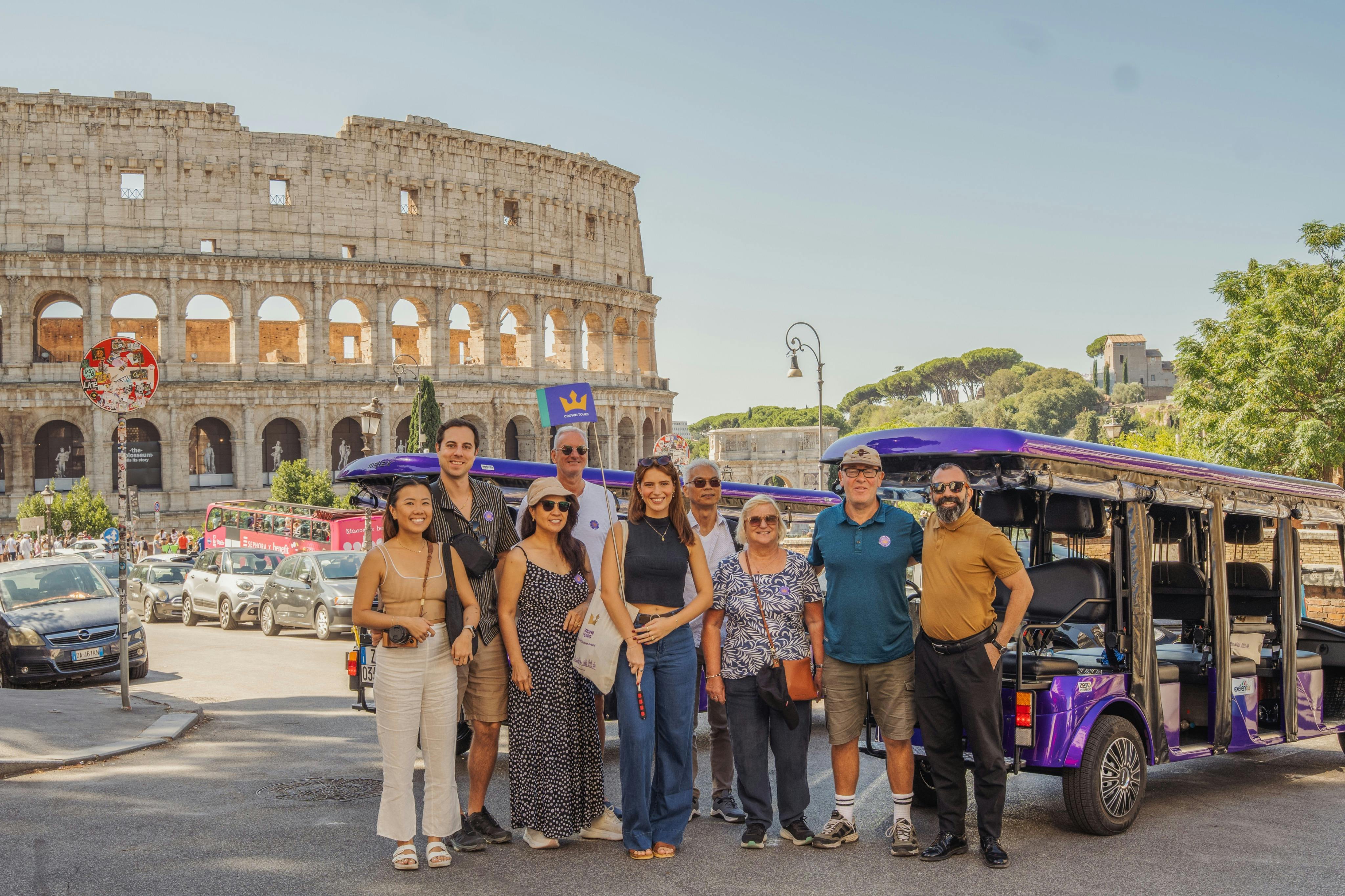 Un gruppo di persone posa davanti al Colosseo a Roma in una giornata di sole, con auto e turisti sullo sfondo.