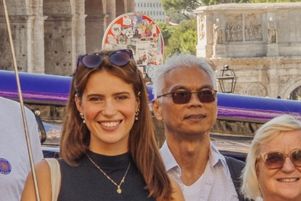 A group of people poses in front of the Colosseum in Rome on a sunny day, with cars and tourists in the background.