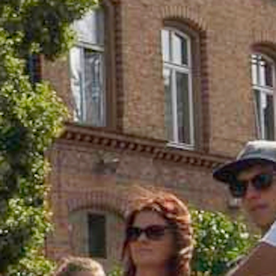 A group of people on bicycles gather outdoors near brick buildings and large trees, engaged in conversation or listening.