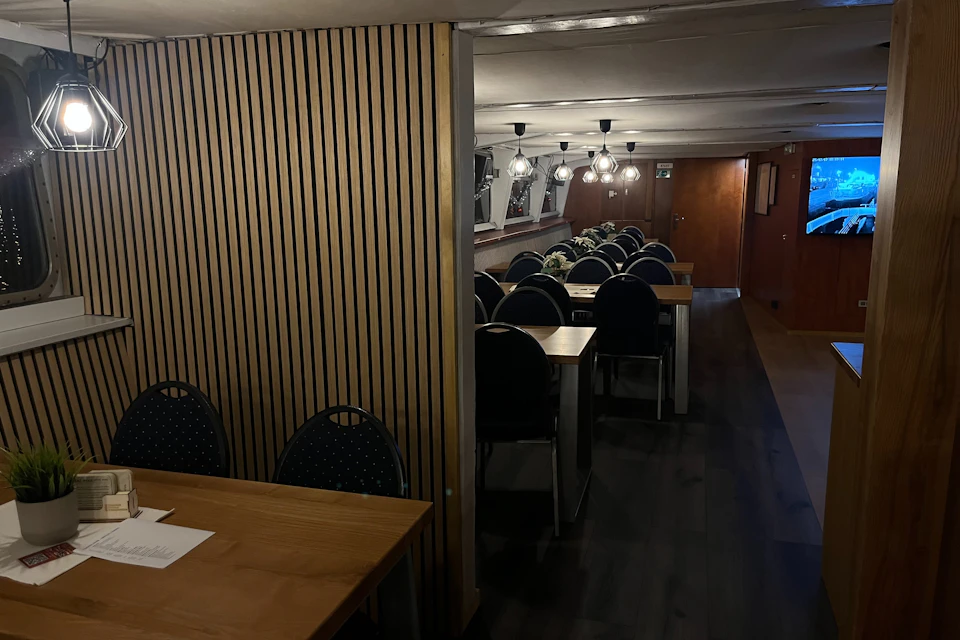 A dimly lit interior with wooden tables and chairs arranged in rows, decorative plants, and hanging light fixtures.
