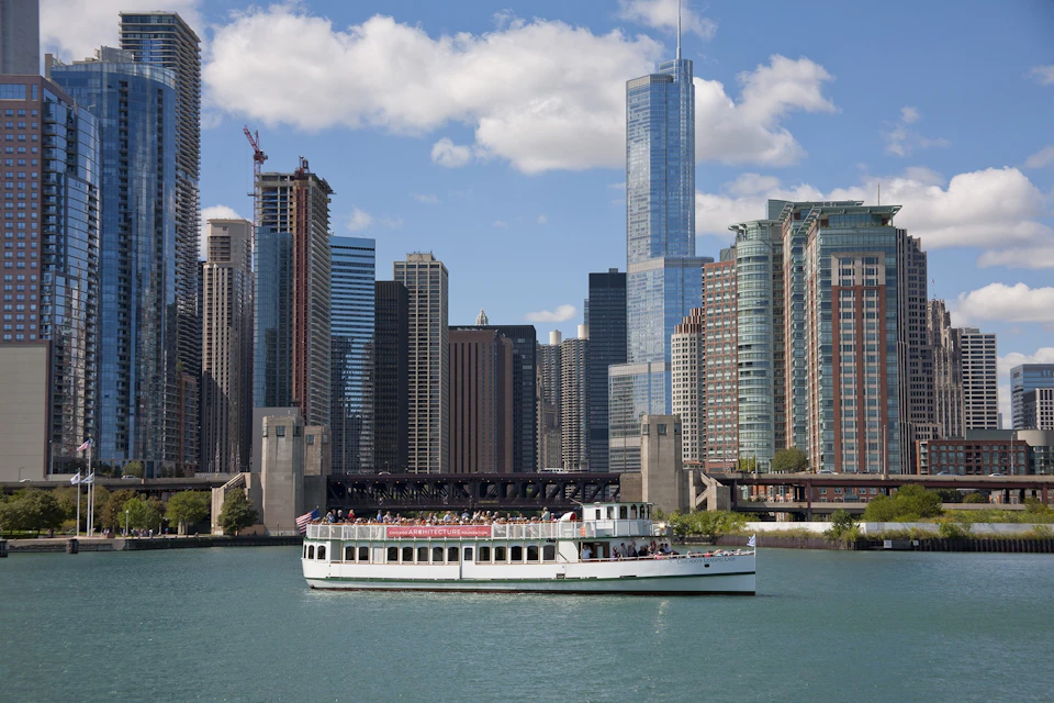 A white tour boat on a river with people aboard in front of a city skyline with modern skyscrapers and a partly cloudy sky.