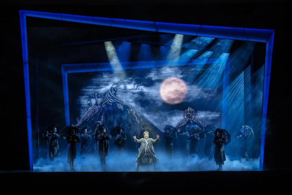 A theatrical performance with a moonlit backdrop, fog effects, and actors in dark costumes on stage under dramatic lighting.