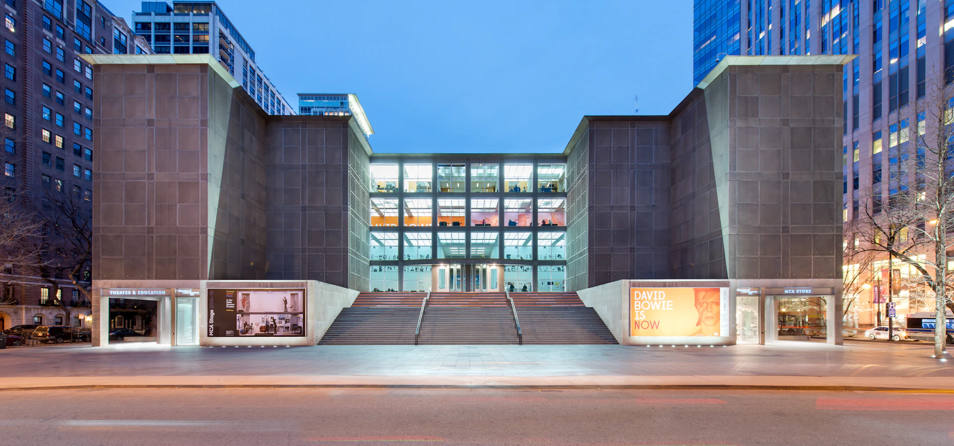 Modern building with large glass windows, central staircase, and "David Bowie Is Now" exhibition banner on the right.