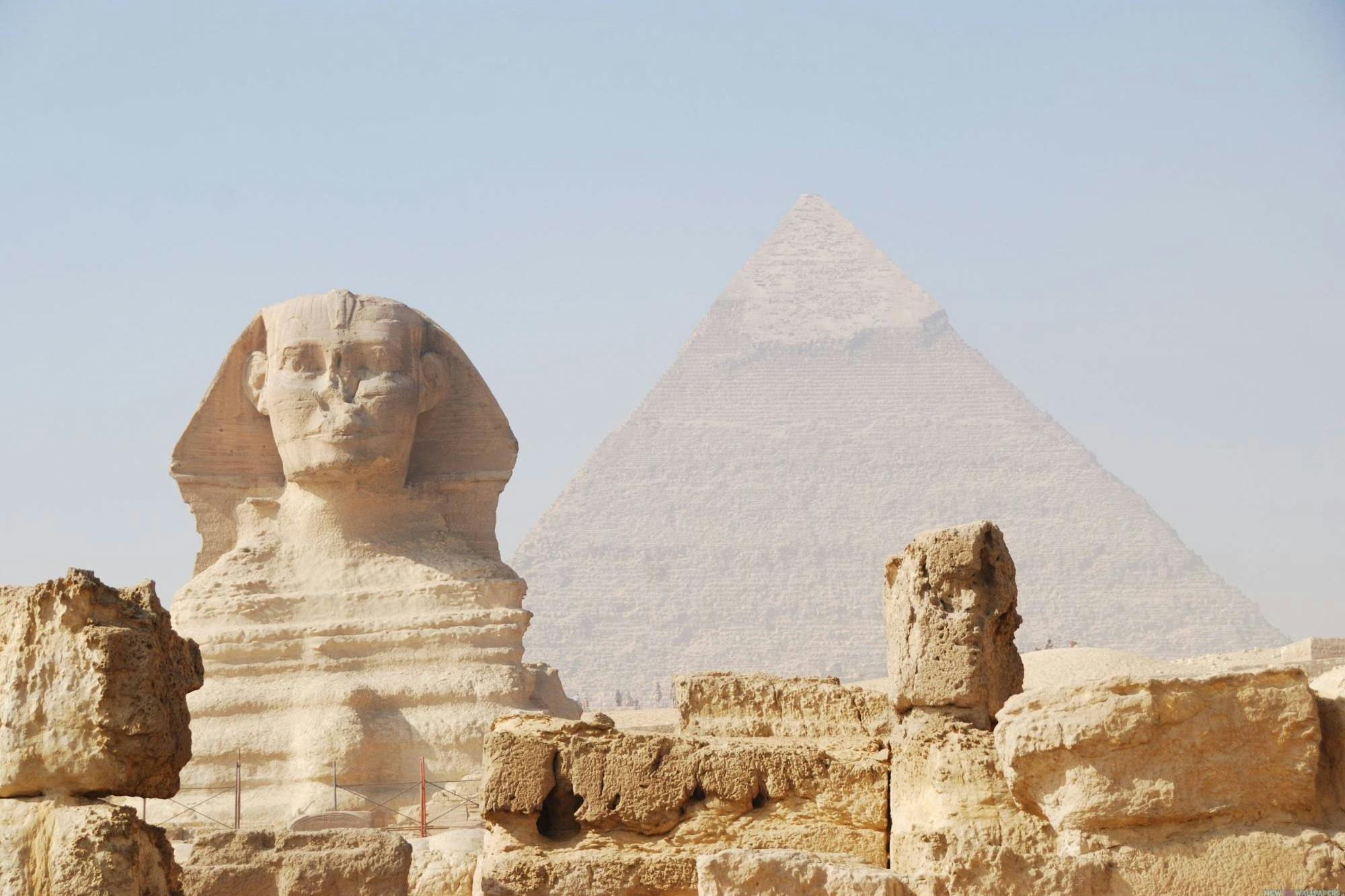 The Great Sphinx of Giza with the Great Pyramid in the background, surrounded by sandy terrain and ancient stone remains.