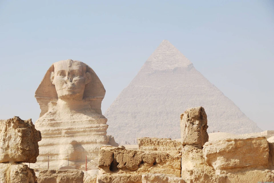 The Great Sphinx of Giza with the Great Pyramid in the background, surrounded by sandy terrain and ancient stone remains.