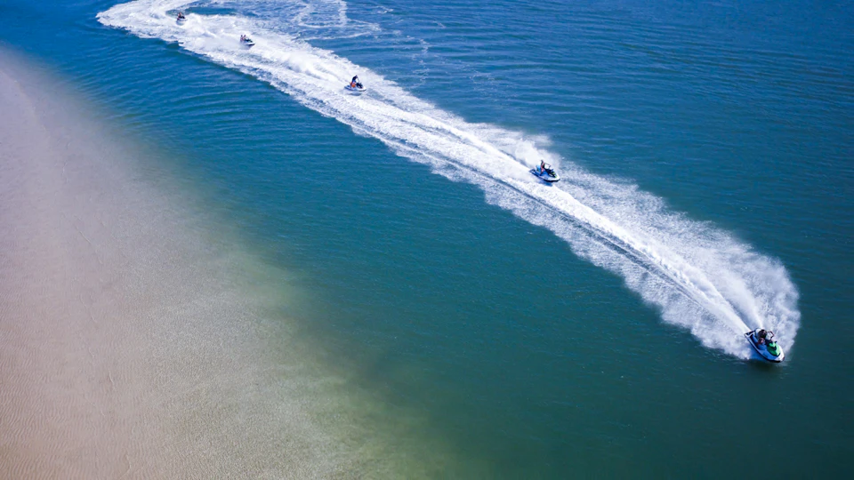 Three jet skis create wake trails as they speed across clear blue water near a sandy shore.