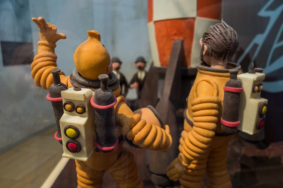 Two figurines in orange spacesuits with jetpacks stand near a rocket model. A group of small figures is visible in the background.
