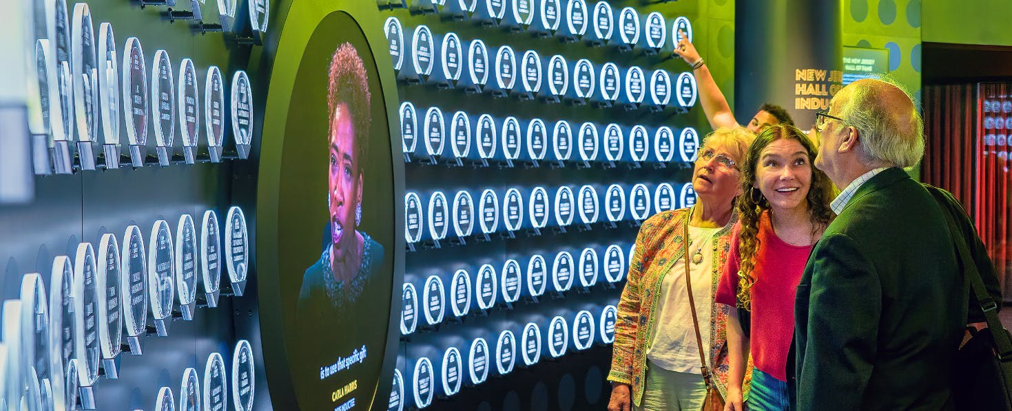 Tři lidé pozorují stěnu s osvětlenými plaketami a kruhovou digitální obrazovkou zobrazující ženský obličej a text.