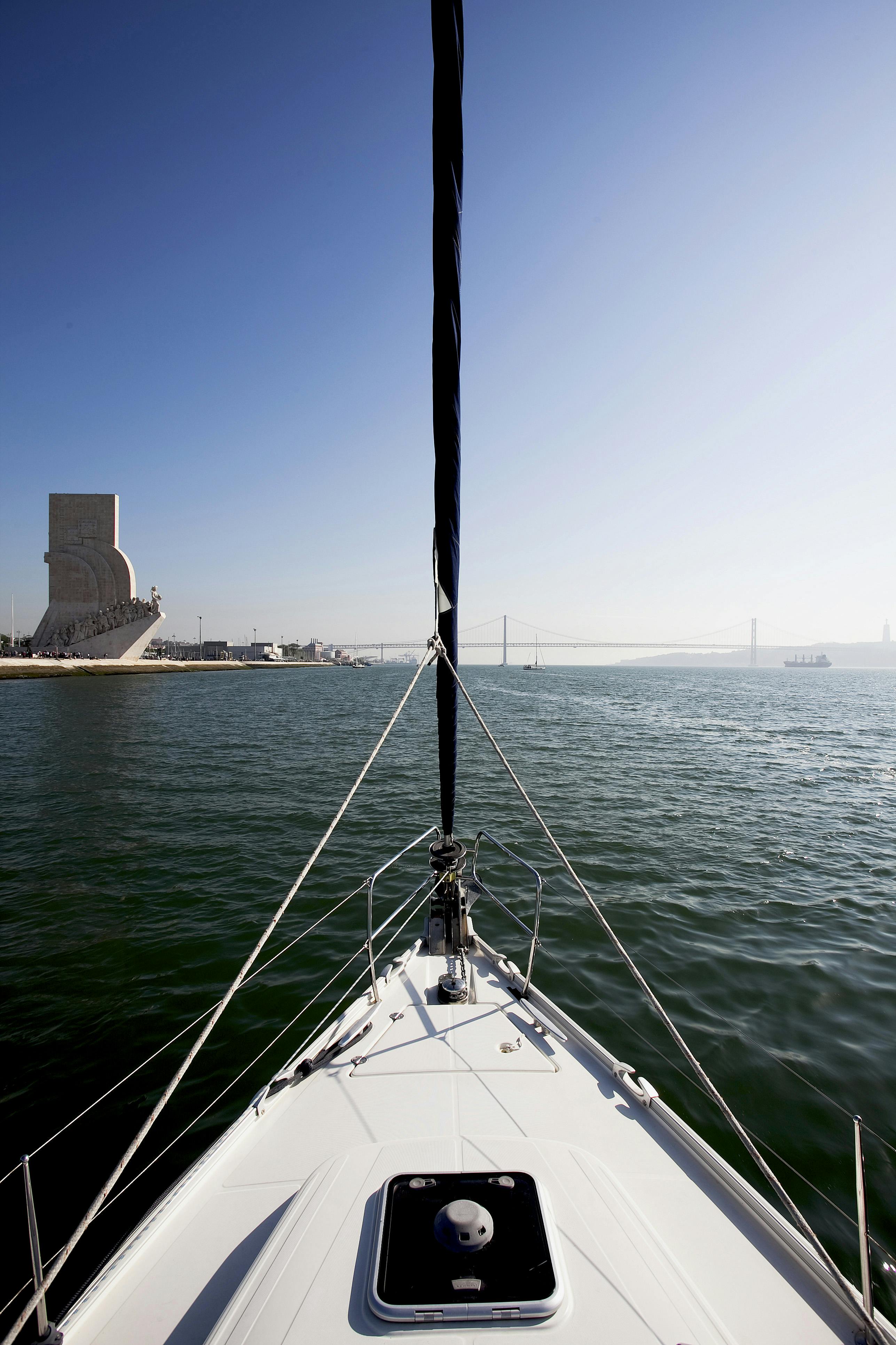 Vista dalla prua di una barca a vela, con un monumento sulla sinistra, un ponte sospeso sullo sfondo e un'ampia distesa d'acqua sotto un cielo limpido.
