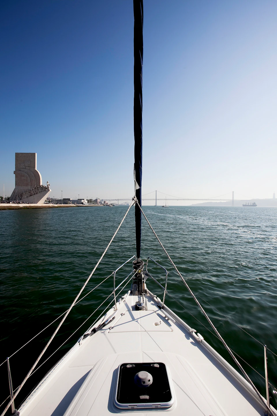 View from the bow of a sailboat, with a monument on the left, a suspension bridge in the background, and expansive water under a clear sky.