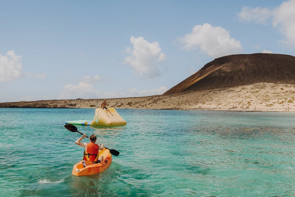 Person kayaking towards an inflatable structure on turquoise water, with a rocky hill and cloudy sky in the background.