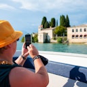 Person in a yellow hat taking a photo with a smartphone from a boat, capturing buildings and trees across a body of water.