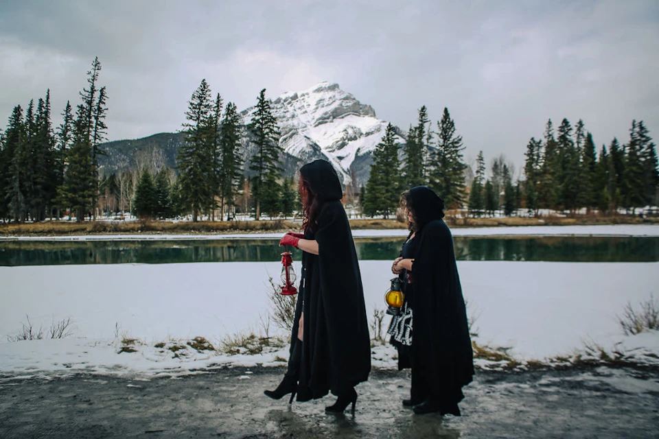 Two people in dark cloaks walk by a snowy river, holding lanterns. Snowy mountain and evergreen trees in the background.