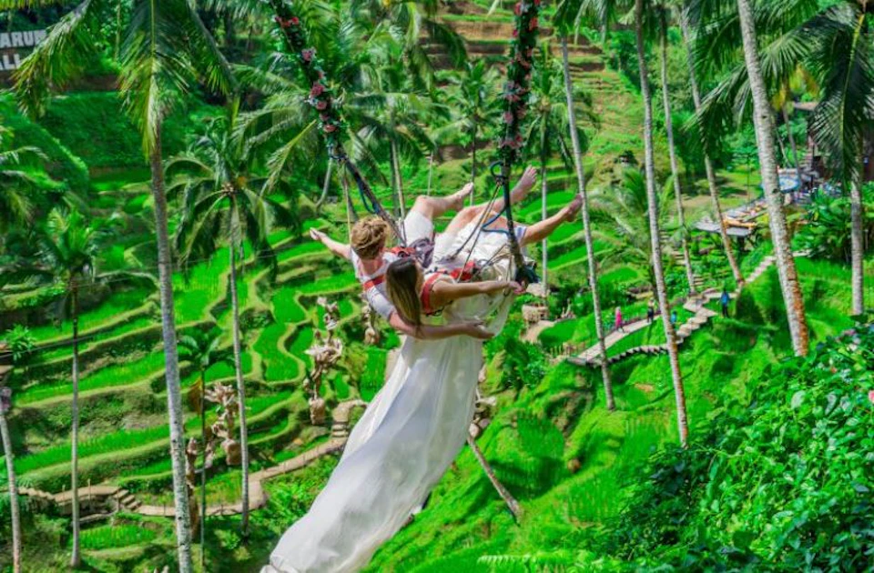 A couple on a swing above vibrant green rice terraces surrounded by tall palm trees. The woman is wearing a long white dress.
