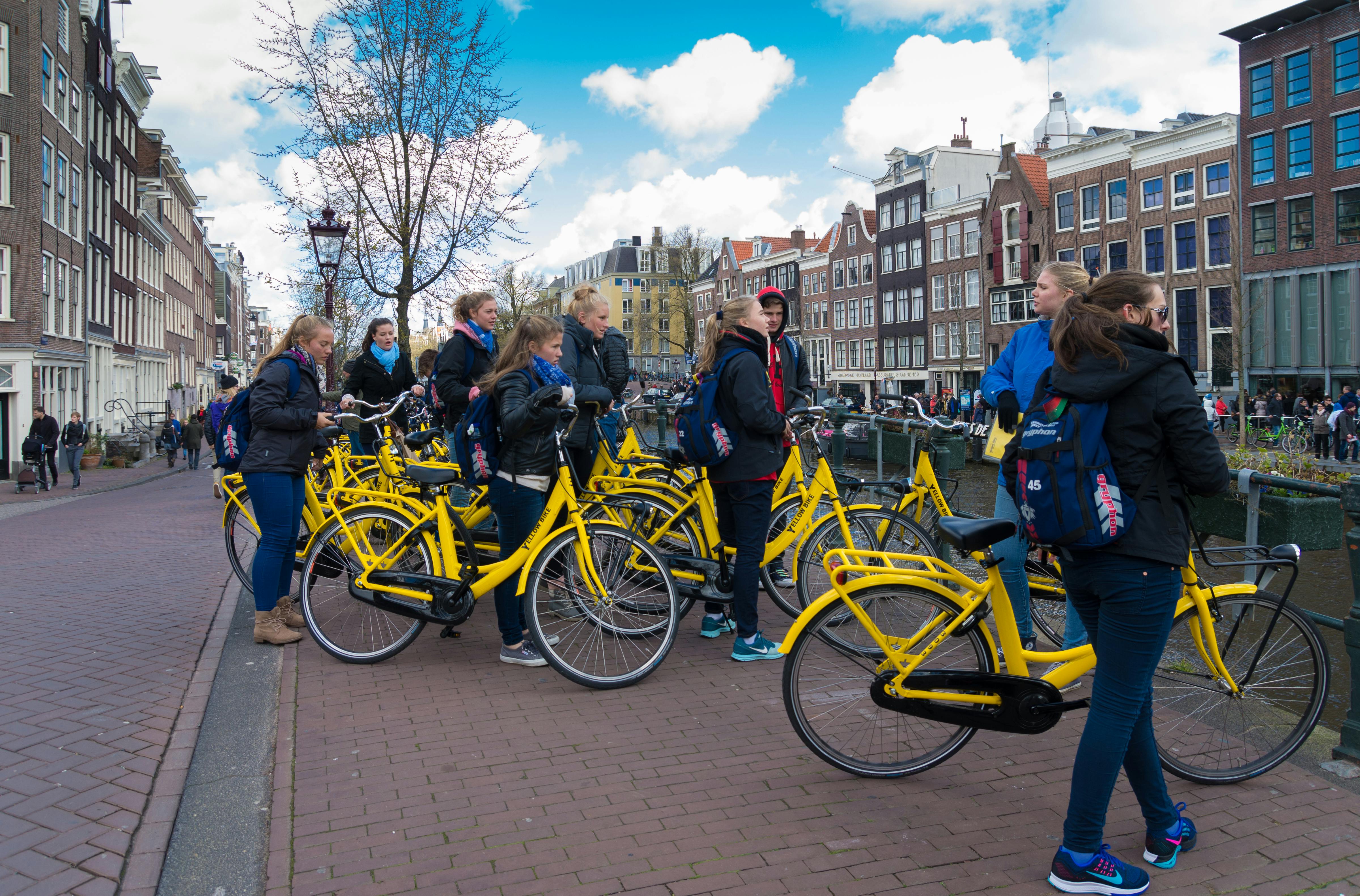 Um grupo de pessoas com mochilas está ao lado de bicicletas amarelas num caminho de calçada perto de um canal e de edifícios históricos sob um céu azul.