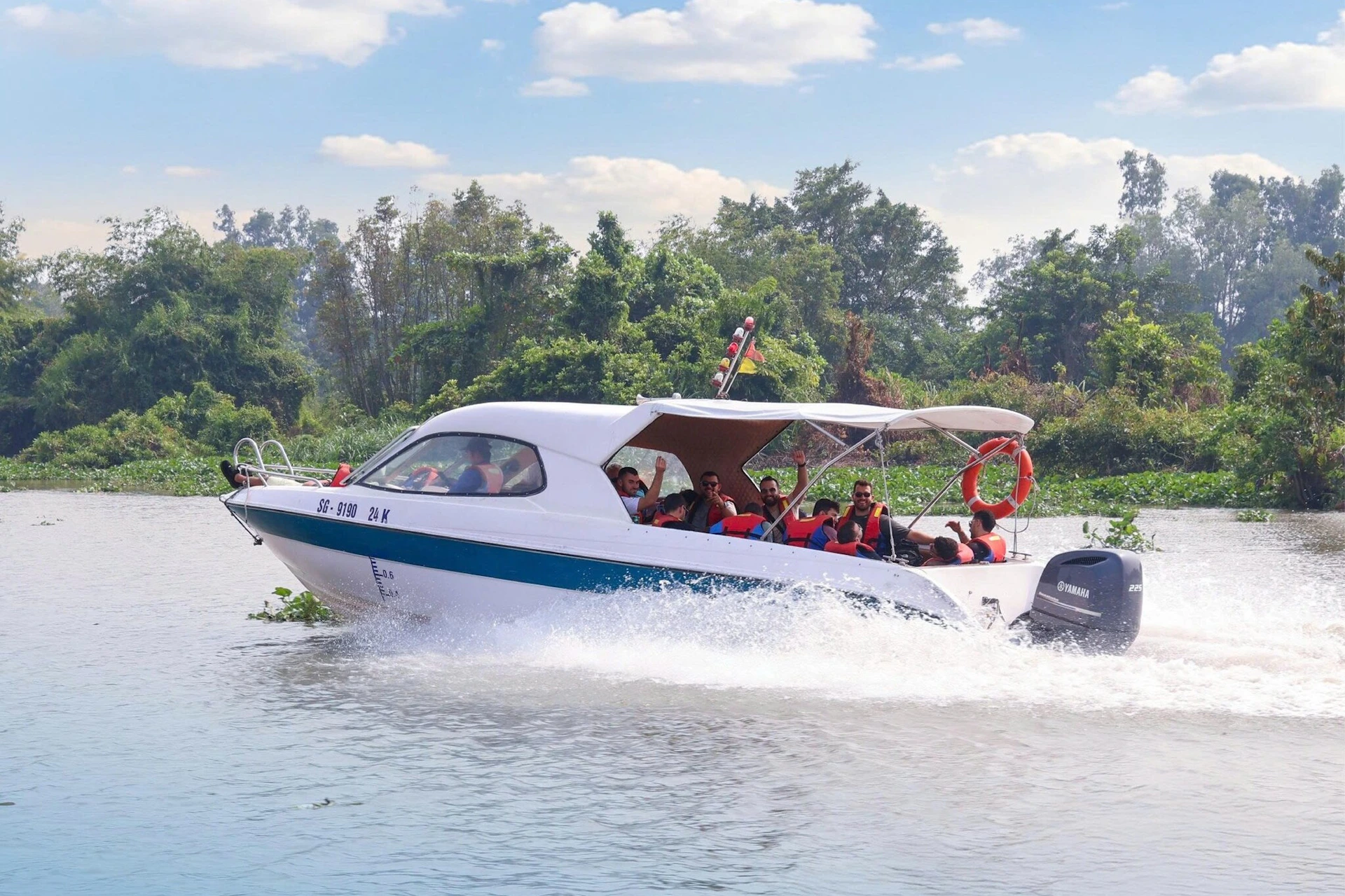 A motorized boat with passengers wearing life jackets cruising on a river, surrounded by greenery and under a blue sky.