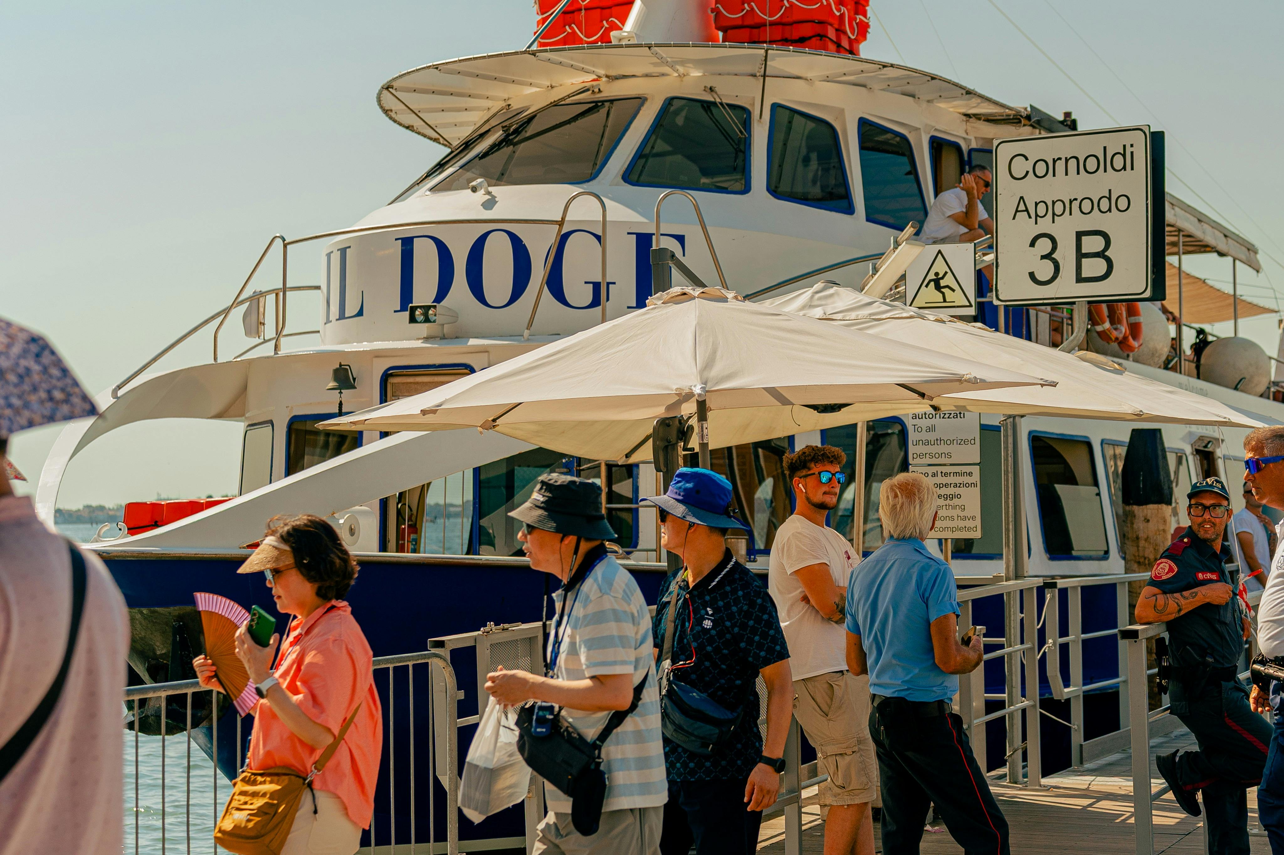 日当たりの良い空の下、「Cornoldi Approdo 3 B」と書かれた看板を掲げたドッキングボートの近くを歩く人々。