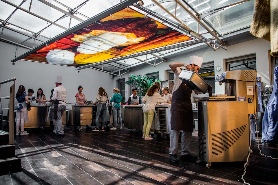 People cooking in a modern kitchen with chefs assisting, under a large colorful food-themed ceiling mural.
