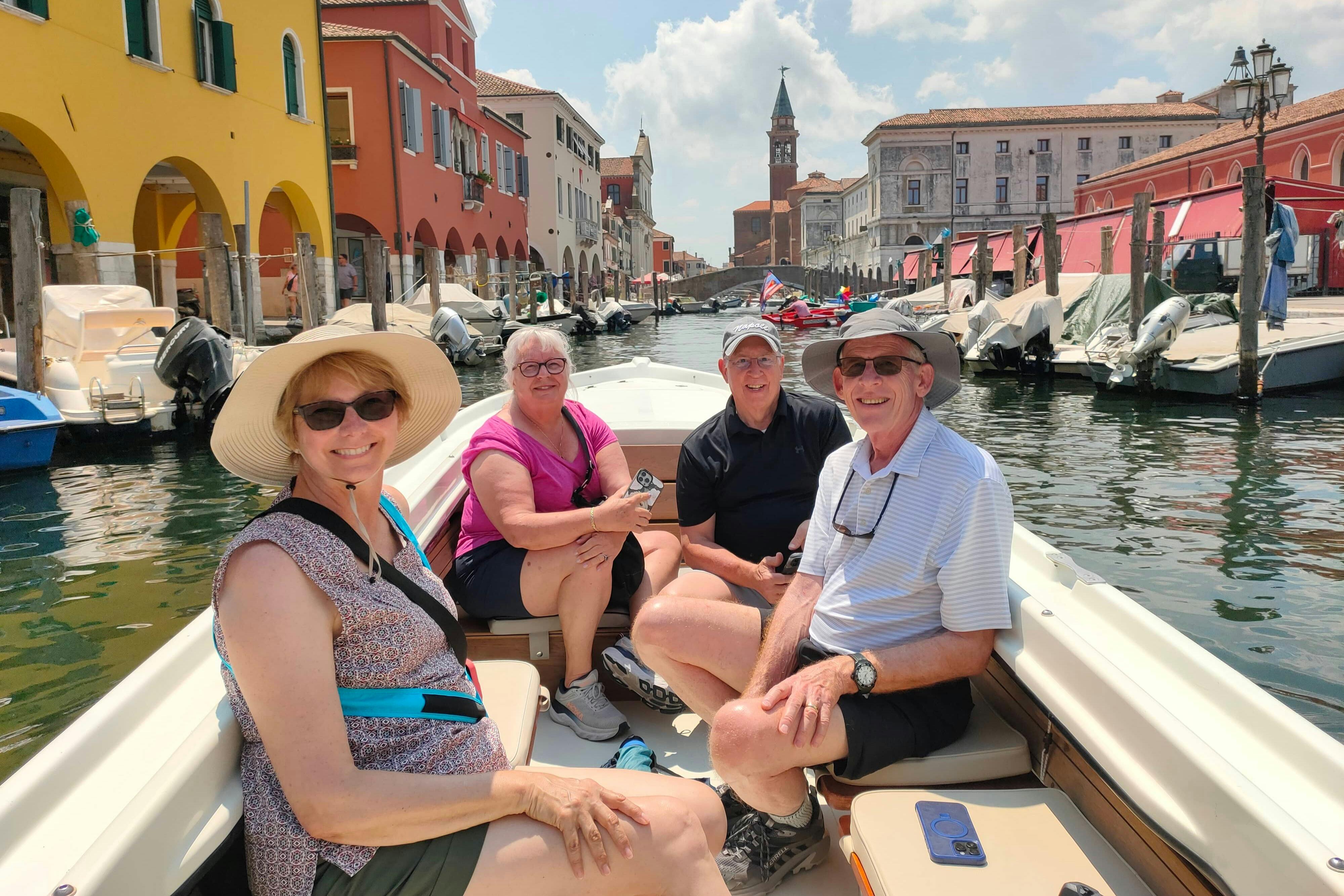 Four people sitting on a boat in a canal surrounded by colorful buildings and boats on a sunny day.