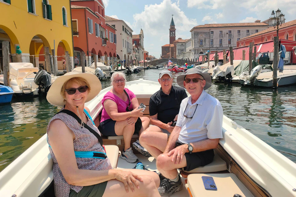 Four people sitting on a boat in a canal surrounded by colorful buildings and boats on a sunny day.