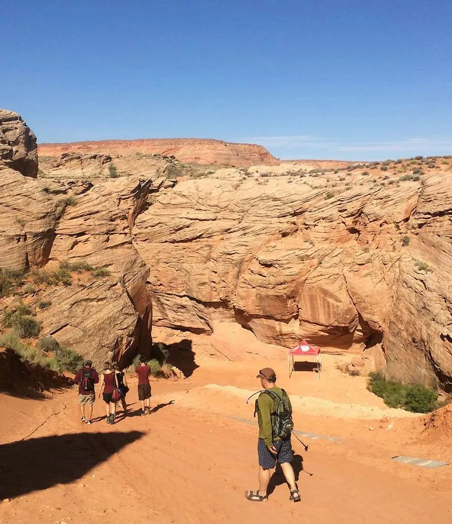 A group of hikers walking towards a rocky canyon under a clear blue sky, with a small red tent in the distance.