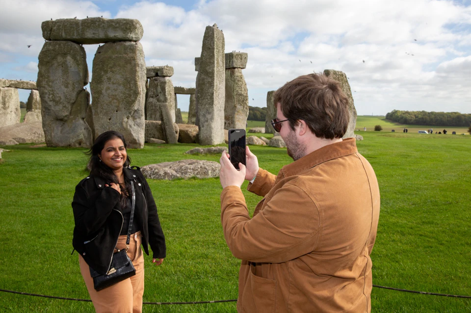 A person smiles while posing for a photo near Stonehenge, with another person holding a smartphone to take the picture.