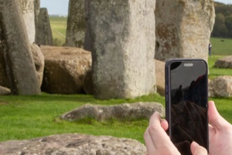 A person smiles while posing for a photo near Stonehenge, with another person holding a smartphone to take the picture.