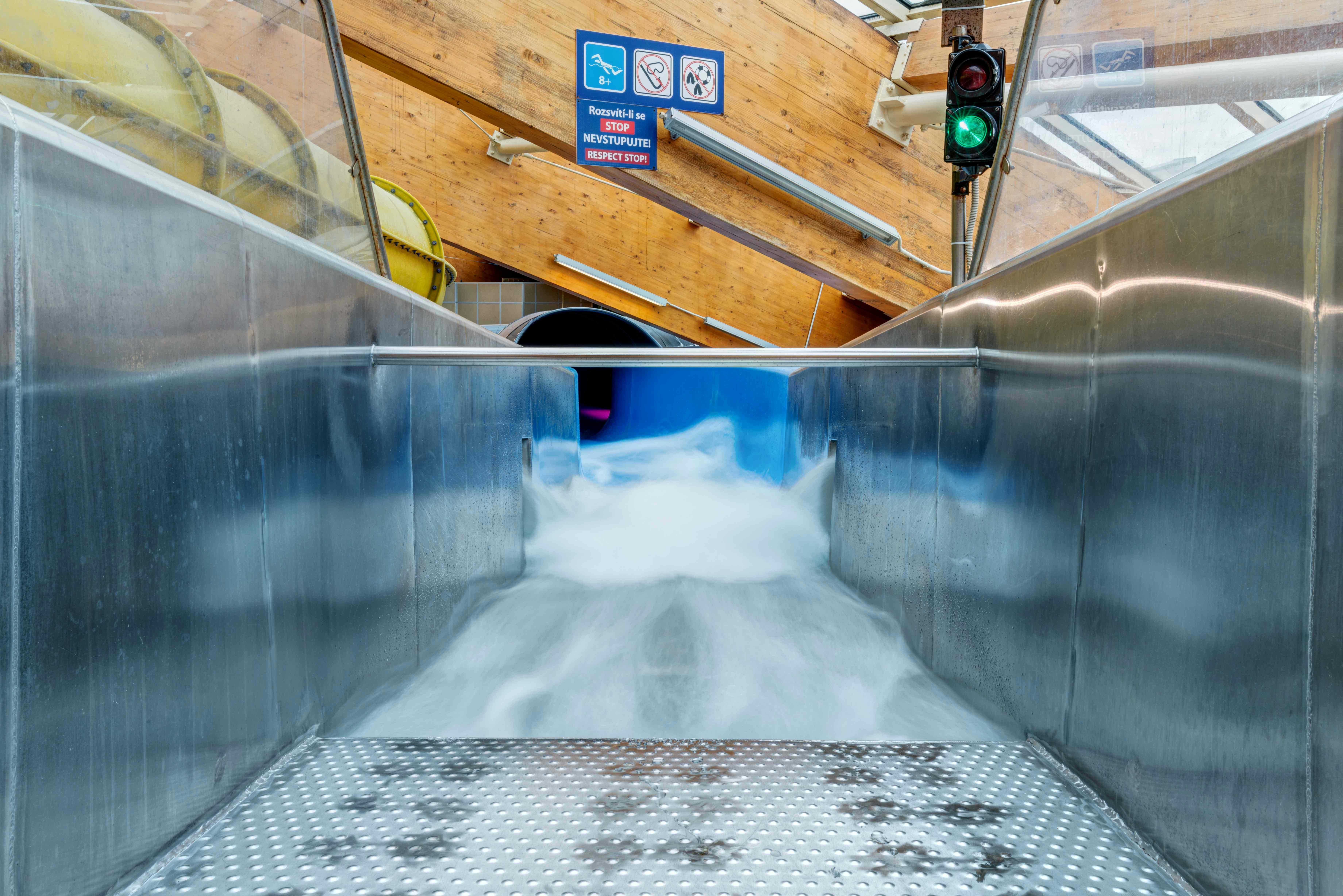 A water slide with flowing water, metal sides, and a "No Entry" sign at the top inside an indoor waterpark facility.