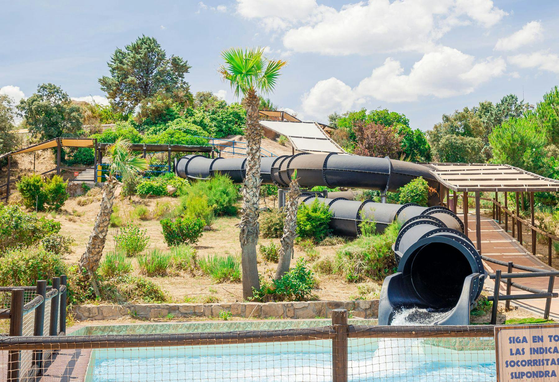 Un parc aquatique avec des toboggans tubulaires noirs, entouré d'arbres et de verdure, sous un ciel partiellement nuageux.