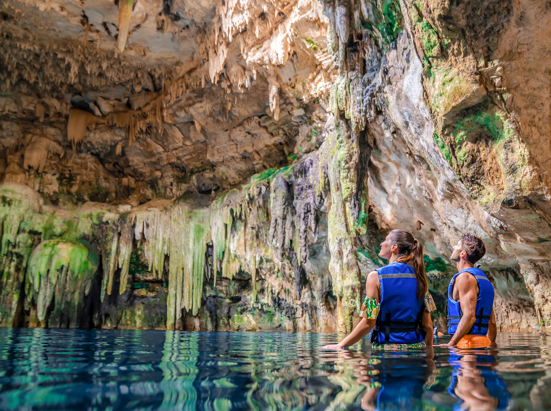 Two people in life jackets stand in water, looking up at stalactites and rock formations inside a cave.