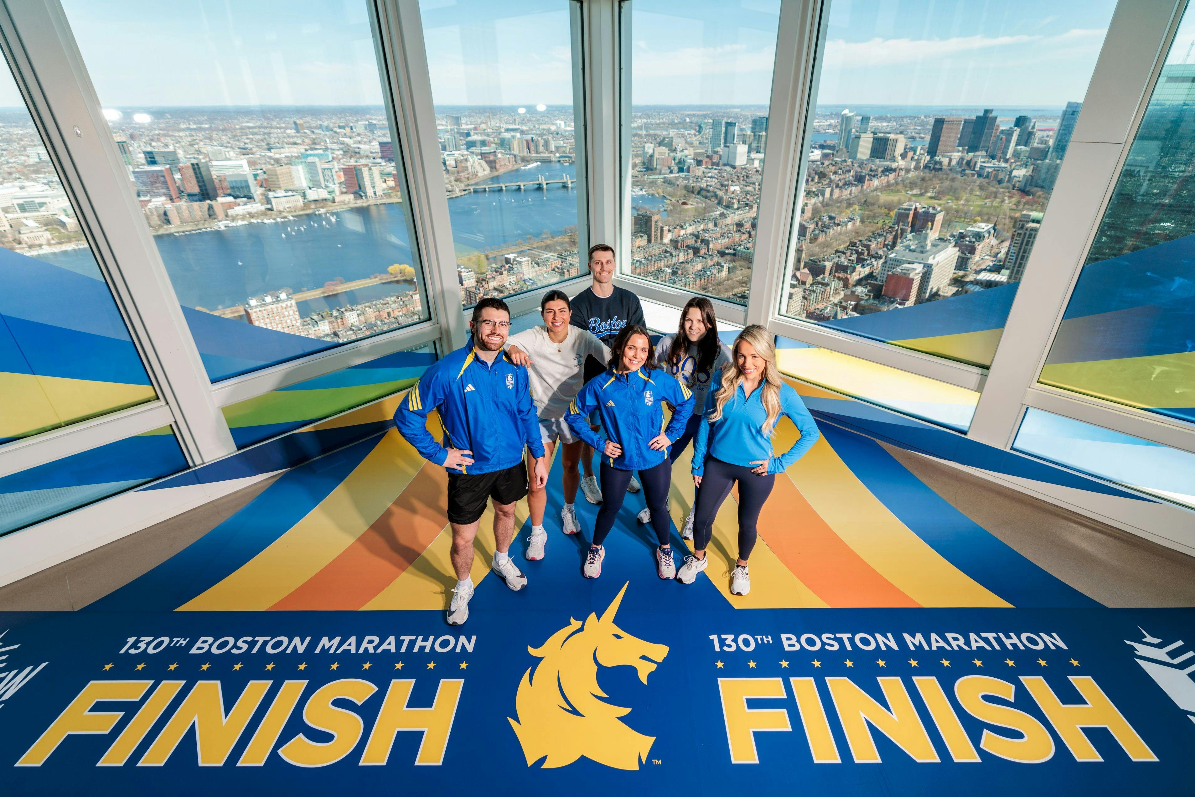 Um grupo de pessoas posando num chão desenhado com gráficos da linha de chegada da "Maratona de Boston", acima de uma vista da cidade.