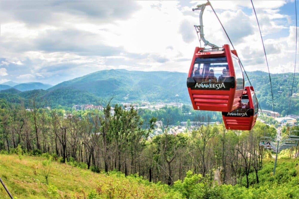 Two red Anakeesta gondolas ride above a forested hillside, with a scenic view of mountains and a town in the background.