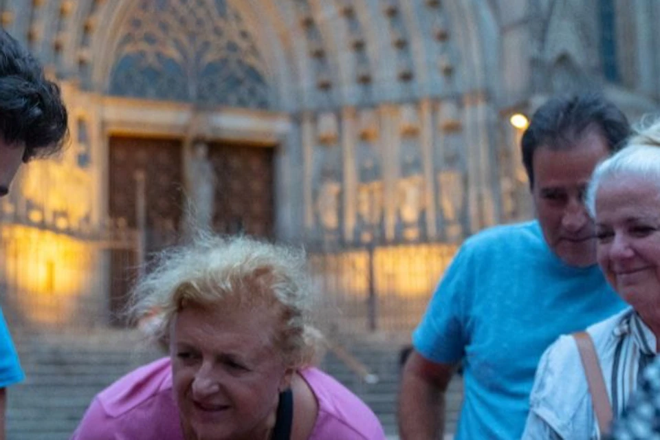 A group of people gathers outside a large, ornate cathedral, looking at something a man in a red shirt is showing them.
