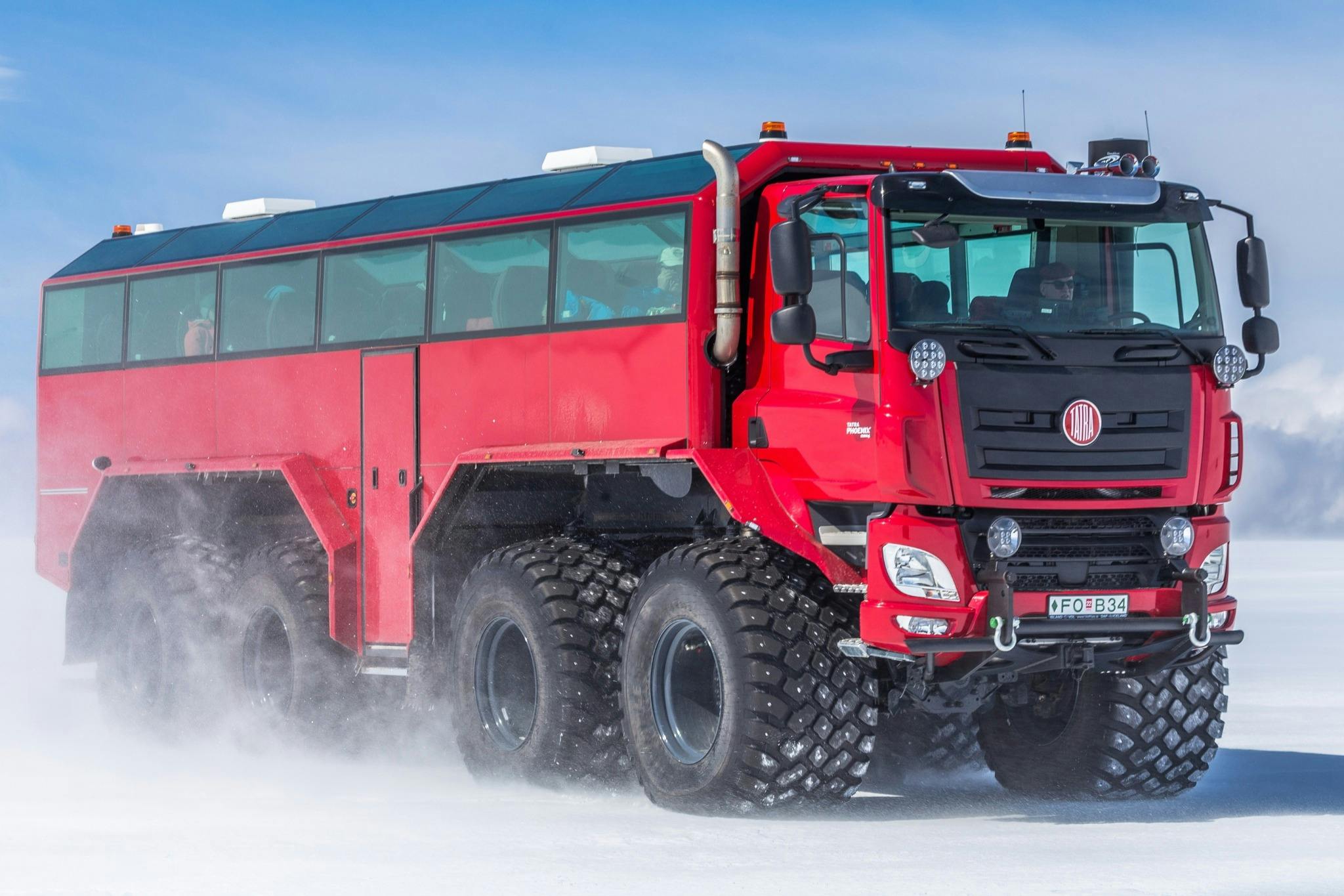 Velké červené terénní vozidlo s více pneumatikami jedoucí po zasněženém terénu pod jasně modrou oblohou.
