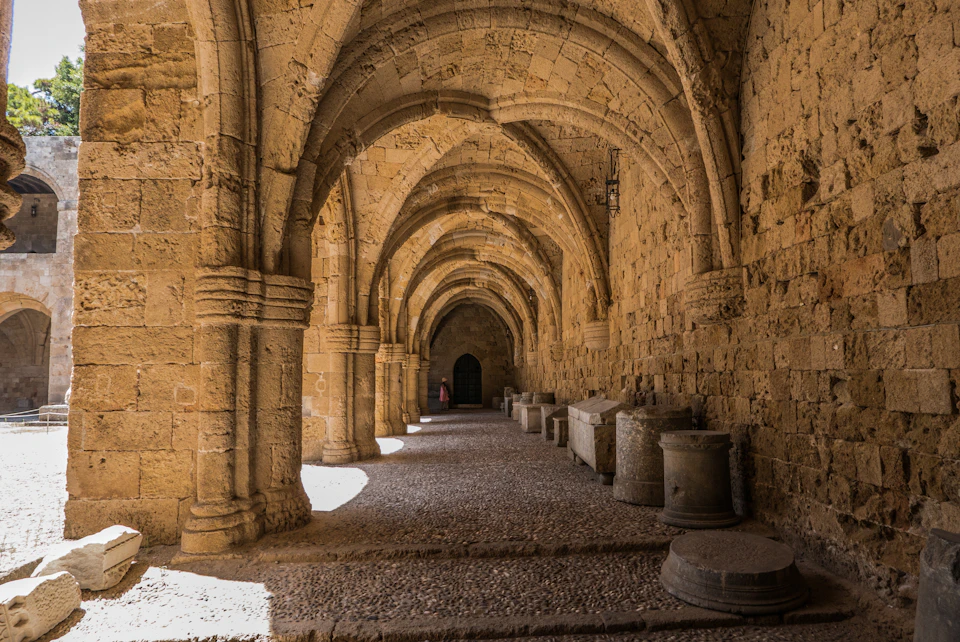 Stone archway corridor with vaulted ceiling, row of ancient tombs, and a distant dark door, lit by natural light from the left.